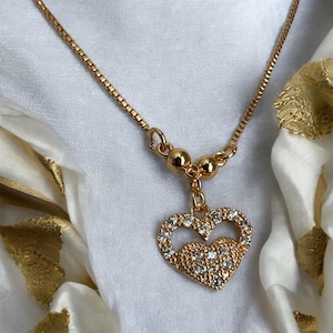 May include: A gold-toned necklace featuring a box chain and a heart-shaped pendant. The pendant consists of an open heart outline and a solid heart base, both encrusted with clear, sparkling stones. Two small gold spheres sit above the pendant.