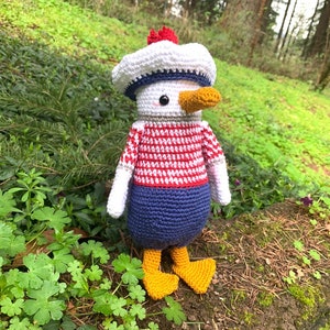 May include: A handmade crochet duck doll wearing a sailor outfit. The doll has a white hat with a blue band and a red pom-pom. The duck has a red and white striped shirt, blue pants, and yellow feet. The doll is standing on a mossy surface.