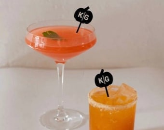 Peut inclure: Une coupe à pied remplie d'un cocktail rose garni d'une feuille verte et d'un pic noir en forme de citrouille marqué KG, à côté d'un verre court contenant un cocktail orange avec des glaçons et un pic noir assorti en forme de citrouille KG.