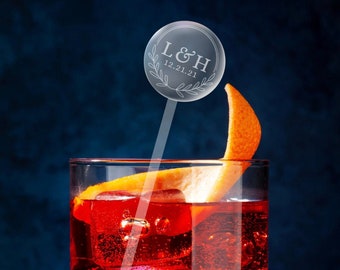 Peut inclure: Un agitateur à cocktail en verre transparent avec un dessus rond en acrylique orné du texte "L&H 12.21.21" dans un design de couronne. L'agitateur est dans un verre rempli d'un cocktail rouge et d'une garniture de zeste d'orange.