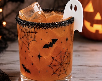 May include: A clear glass tumbler filled with an orange liquid and ice cubes, featuring a black rim of sugar and black printed graphics of spiderwebs, bats, and stars. A white, flat, ghost-shaped garnish is attached to the rim of the glass.