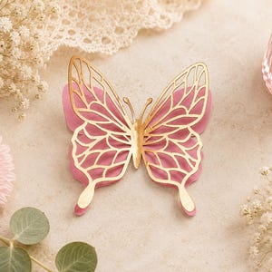 Pode incluir: Um ornamento decorativo de borboleta com um design em camadas, apresentando uma base rosa s&oacute;lida e uma camada superior met&aacute;lica dourada com padr&otilde;es recortados intrincados nas asas. A borboleta tem uma forma sim&eacute;trica e antenas detalhadas.