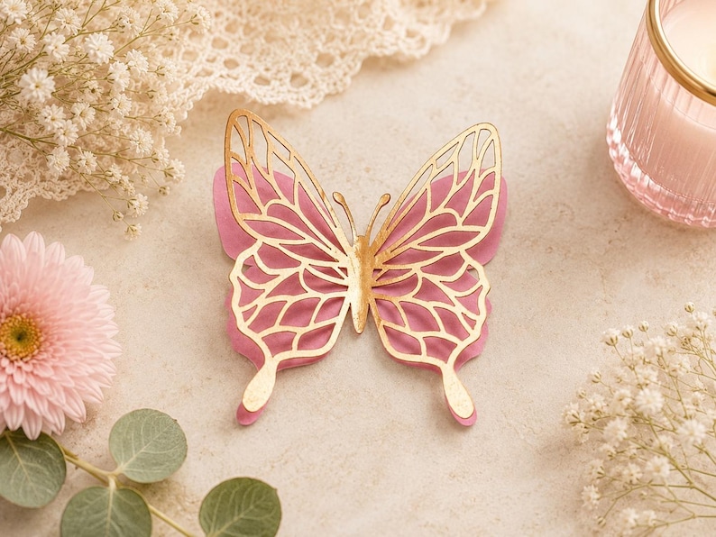 Pode incluir: Um ornamento decorativo de borboleta com um design em camadas, apresentando uma base rosa s&oacute;lida e uma camada superior met&aacute;lica dourada com padr&otilde;es recortados intrincados nas asas. A borboleta tem uma forma sim&eacute;trica e antenas detalhadas.
