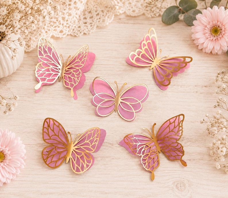 Pode incluir: Cinco recortes decorativos de borboletas com um design em camadas, apresentando asas de papel rosa s&oacute;lido por baixo e sobreposi&ccedil;&otilde;es de filigrana met&aacute;lica dourada. Cada borboleta exibe uma forma de asa e um padr&atilde;o &uacute;nicos e distintos.