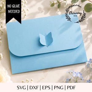 Puede incluir: Un sobre de papel azul claro con una pestaña de cierre troquelada en forma de cabeza de gato. El texto NO GLUE NEEDED aparece en una pancarta negra arriba a la izquierda, y el texto Dreaming SVG se muestra en un diseño de corona de laurel.