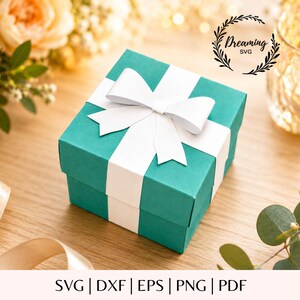 Può includere: Una scatola regalo quadrata color verde acqua con un nastro bianco annodato a fiocco sulla parte superiore. La scatola è centrata e il testo Dreaming SVG è visibile nell'angolo in alto a destra. In basso appare il testo SVG | DXF | EPS | PNG | PDF.