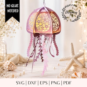 May include: A hanging paper jellyfish lantern in shades of light purple and pink, featuring intricate floral cutouts and dangling tentacle strips. The structure is illuminated from within, and the text NO GLUE NEEDED and Dreaming SVG are visible.