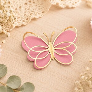 Pode incluir: Uma decora&ccedil;&atilde;o em forma de borboleta com um design em camadas, apresentando uma base rosa s&oacute;lida e uma camada superior met&aacute;lica dourada com detalhes de asas recortadas e duas antenas finas que se estendem do centro do corpo.