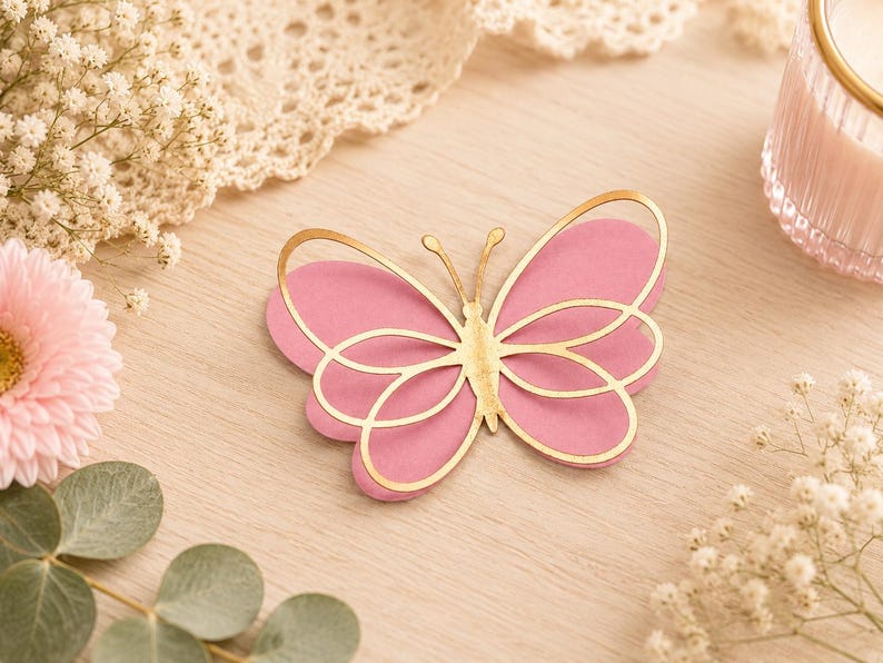 Pode incluir: Uma decora&ccedil;&atilde;o em forma de borboleta com um design em camadas, apresentando uma base rosa s&oacute;lida e uma camada superior met&aacute;lica dourada com detalhes de asas recortadas e duas antenas finas que se estendem do centro do corpo.