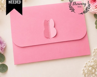 May include: A pink paper envelope featuring a die-cut rabbit ear closure mechanism on the front flap. The text NO GLUE NEEDED appears in a black banner in the upper left corner, while the text Dreaming SVG is enclosed in a circular wreath.