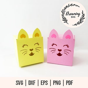 Può includere: Due piccole scatole di cartone vuote a forma di gatto. La scatola a sinistra è gialla con il naso rosa e i baffi. La scatola a destra è rosa con il naso marrone e i baffi. Entrambe le scatole hanno orecchie a forma di cuore.