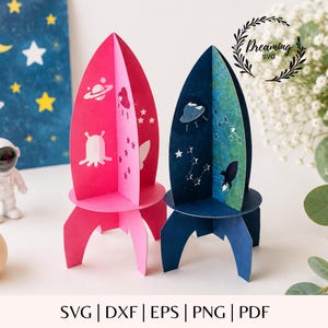May include: Two 3D rocket ship paper crafts, one in bright pink and one in dark blue with a green gradient. The pink rocket features white space-themed icons, while the blue rocket displays white constellations and a small rocket silhouette.
