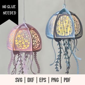 May include: Two papercraft jellyfish lanterns, one blue and one pink, with intricate floral designs and dangling tentacles. The lanterns are lit from within, creating a warm glow. The text "NO GLUE NEEDED" is visible in the top left corner.