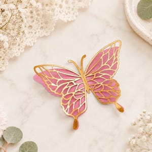 Pode incluir: Um ornamento decorativo de borboleta com um design em camadas, apresentando uma base rosa s&oacute;lida e uma camada superior de filigrana dourada met&aacute;lica. O intrincado padr&atilde;o recortado revela a cor rosa por baixo, criando um contraste entre o dourado e o rosa.