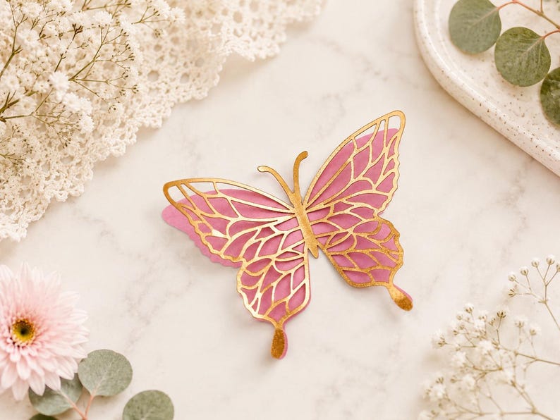 Pode incluir: Um ornamento decorativo de borboleta com um design em camadas, apresentando uma base rosa s&oacute;lida e uma camada superior de filigrana dourada met&aacute;lica. O intrincado padr&atilde;o recortado revela a cor rosa por baixo, criando um contraste entre o dourado e o rosa.