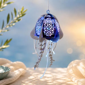 May include: A hanging jellyfish ornament featuring a layered structure with a deep blue, intricate geometric cut-out pattern on the bell. The piece includes multiple dangling, thin, light-colored tentacles that extend downward from the main body.