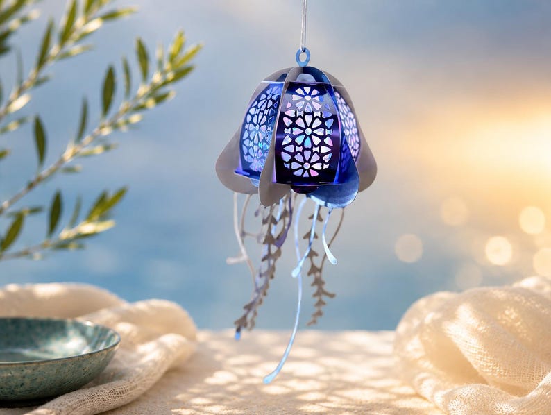 May include: A hanging jellyfish ornament featuring a layered structure with a deep blue, intricate geometric cut-out pattern on the bell. The piece includes multiple dangling, thin, light-colored tentacles that extend downward from the main body.