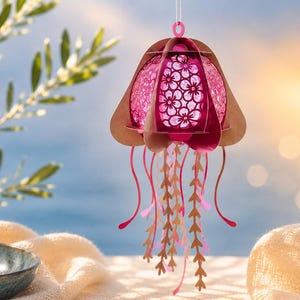 May include: A hanging jellyfish ornament crafted from layered paper in shades of pink and tan. The central dome features a delicate floral cutout pattern, while the lower section consists of multiple dangling, slender, and wavy paper tentacles.