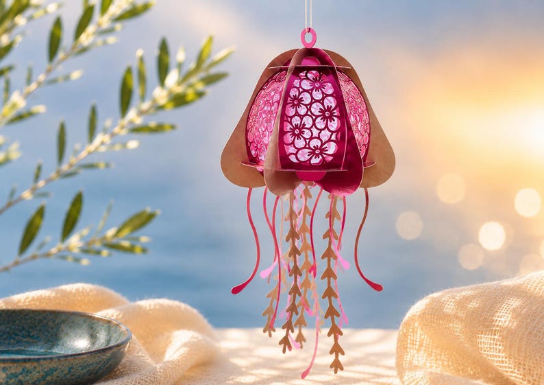 May include: A hanging jellyfish ornament crafted from layered paper in shades of pink and tan. The central dome features a delicate floral cutout pattern, while the lower section consists of multiple dangling, slender, and wavy paper tentacles.