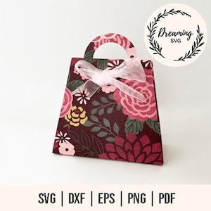 May include: A printable template for a floral gift bag with a pink bow. The bag is designed to be cut out and assembled. The design features a burgundy background with pink, yellow, and green floral patterns. The text "Dreaming SVG" is included in a circle.