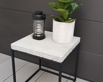 May include: A small concrete side table with a black metal frame. The table is sitting on a gray deck and has a potted plant and a lantern on top.