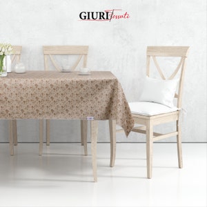 Pode incluir: Uma toalha de mesa bege com padrão floral, colocada em uma mesa de madeira com quatro cadeiras. A toalha de mesa tem uma borda branca e as cadeiras são de madeira clara.
