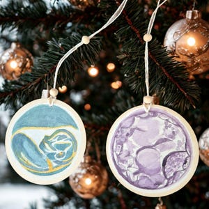 Puede incluir: Dos adornos navideños redondos de madera con diseños de acuarela. Uno presenta remolinos azules y amarillos, el otro un patrón floral púrpura. Cada uno cuelga de una cuerda blanca con una cuenta de madera, sobre un fondo borroso de árbol de Navidad.