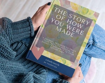 May include: A hardcover book titled "THE STORY OF HOW YOU WERE MADE" with a watercolor design. The book is held in a person's lap, with the text "A JOURNAL DESIGNED TO TRACK YOUR FERTILITY PROCESS, IN ORDER TO EXPLAIN IT TO YOUR FUTURE BABY" below the title.