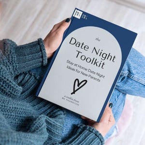 Może przedstawiać: Książka zatytułowana "The Date Night Toolkit" z granatową okładką i białym tekstem. Książka zawiera tekst "Stay at Home Date Night Ideas for New Parents" i symbol serca. Książka jest trzymana przez osobę ubraną w niebieski sweter i dżinsy.