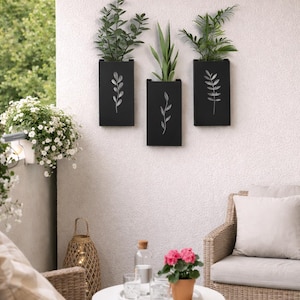 Op de afbeelding: Drie zwarte rechthoekige plantenbakken met witte bladeren hangen aan een witte muur met textuur. Elke plantenbak bevat groene planten. De scène omvat een terras met rotan meubels, een kleine tafel en een roze bloem in een pot.