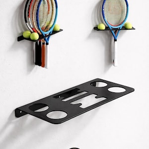 May include: Black metal tennis racket and ball storage shelves mounted on a white wall. The shelves hold tennis rackets and tennis balls. The bottom shelf has cutouts for storage. The logo of a circle with the text "3 Art" is at the bottom.