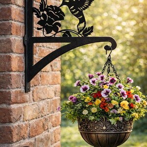 Könnte beinhalten: Schwarzer Metall-Hängekorbhalter mit einem Kolibri- und Blumenmuster. Der Halter trägt einen bronzefarbenen Hängekorb, der mit bunten Blumen gefüllt ist. Der Halter ist an einer Ziegelmauer befestigt.