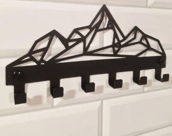 May include: Black metal wall-mounted key holder with a geometric mountain range design. Features five hooks for hanging keys, coats, or other items. The key holder is mounted on a white tiled wall.