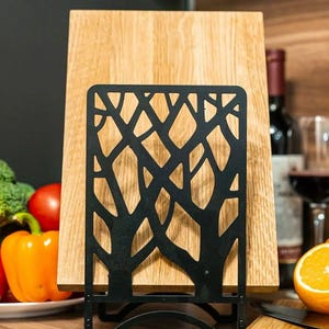 May include: Black metal cutting board stand with a tree branch design. The stand is holding a wooden cutting board. The stand is on a wooden countertop. The text "Art 3 Metal" is visible in the bottom right corner of the image.