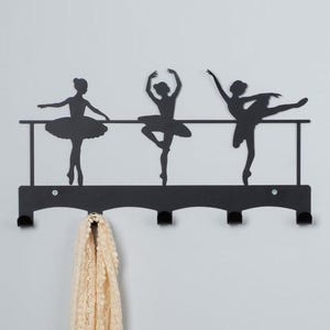 May include: Black metal wall-mounted coat rack featuring a silhouette of three ballerinas in a row. The rack has six hooks and a decorative top.