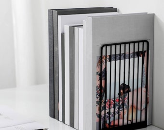 May include: A black metal bookend with a grid design holds a stack of gray and white books. The bookend is on a white surface.