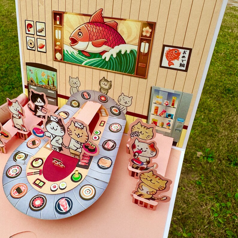 Puede incluir: Una tarjeta desplegable que representa una escena de restaurante de sushi con gatos. La tarjeta est&aacute; abierta y muestra una barra de sushi circular con gatos sentados en el mostrador comiendo sushi. La tarjeta tambi&eacute;n presenta una ilustraci&oacute;n de un pez rojo y el texto "おすし屋" en japon&eacute;s.