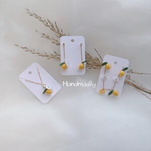 May include: Handmade lemon-themed jewellery set, comprising a necklace and two pairs of earrings, presented on white cards. The pieces feature yellow lemons with green leaves and white flower details, on gold-tone chains. The brand name "Handmadify" is visible.