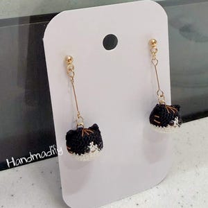 May include: A pair of handmade crochet cat earrings. Each earring features a black and white cat head with gold-colored accents and a long, slender gold-colored drop. The earrings are displayed on a white card.