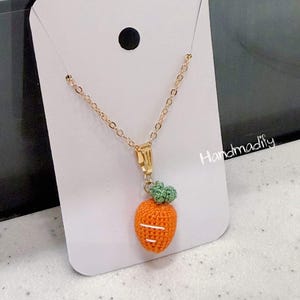 Puede incluir: Collar de cadena dorada con un colgante de zanahoria naranja tejido a ganchillo. La zanahoria tiene hojas verdes y detalles de costura blancos. El collar se muestra en una tarjeta blanca.
