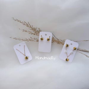 May include: A selection of bee-themed jewellery, including a necklace, earrings, and a floral pendant. The jewellery features yellow and black striped bee designs. The word "Handmadify" is printed on the white background.