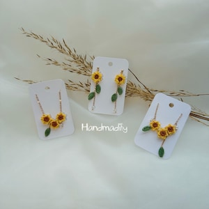 Op de afbeelding: Een set handgemaakte gehaakte sieraden met een zonnebloemthema. De set bevat een ketting en oorbellen, elk met gele zonnebloemen, groene bladeren en goudkleurige kettingen. De tekst "Handmodify" is zichtbaar.