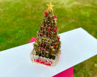 May include: A pop-up Christmas card featuring a gold-colored Christmas tree with red ornaments, bows, and a star on top. The card includes the text "Merry Christmas" and is set against a white and red background.