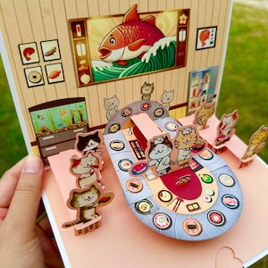 Puede incluir: Una tarjeta pop-up que representa un restaurante de sushi con un pez rojo y una barra de sushi donde los gatos comen sushi. La tarjeta est&aacute; hecha de papel y tiene un efecto 3D.