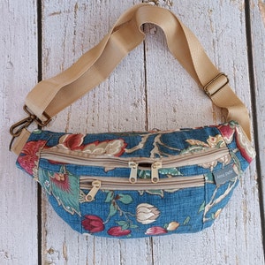 May include: A blue floral fanny pack with a beige adjustable strap and three zippered compartments. The bag features a pattern of red, green, and beige flowers and foliage. The bag has a small tag that says "Anna Sheffe".