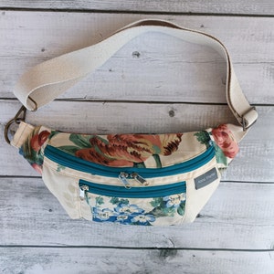 May include: A white canvas fanny pack with a floral print and teal zipper accents. The bag has two zippered compartments and a small label that reads "Anna Shaffer".