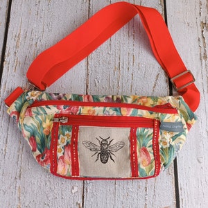 May include: A red and white floral fanny pack with a zippered pocket and a bee design. The fanny pack has a red adjustable strap and the text "Anna Sheffield" is printed on the side.