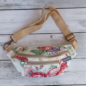 May include: A beige floral fabric fanny pack with two zippered compartments and a brown adjustable strap. The strap has a metal clasp and the bag has a label that reads "Anna Shaffer".