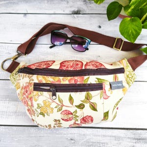 Puede incluir: Un bolso de cintura crema con un estampado floral de granadas rojas y hojas verdes. El bolso tiene una correa y cremalleras marrones, y un par de gafas de sol descansan encima. El bolso tiene el texto "Anna Sheffer".