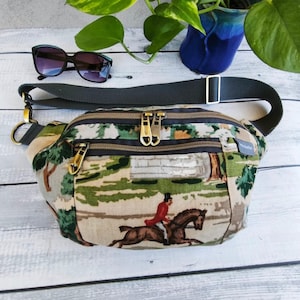 以下が含まれることがあります： 馬に乗ったライダーが描かれた風景プリントのファニーパック。バッグには、ゴールドカラーの金具が付いた複数のジッパー付きコンパートメントがあります。調節可能なストラップは濃い緑色で、サングラスも近くにあります。